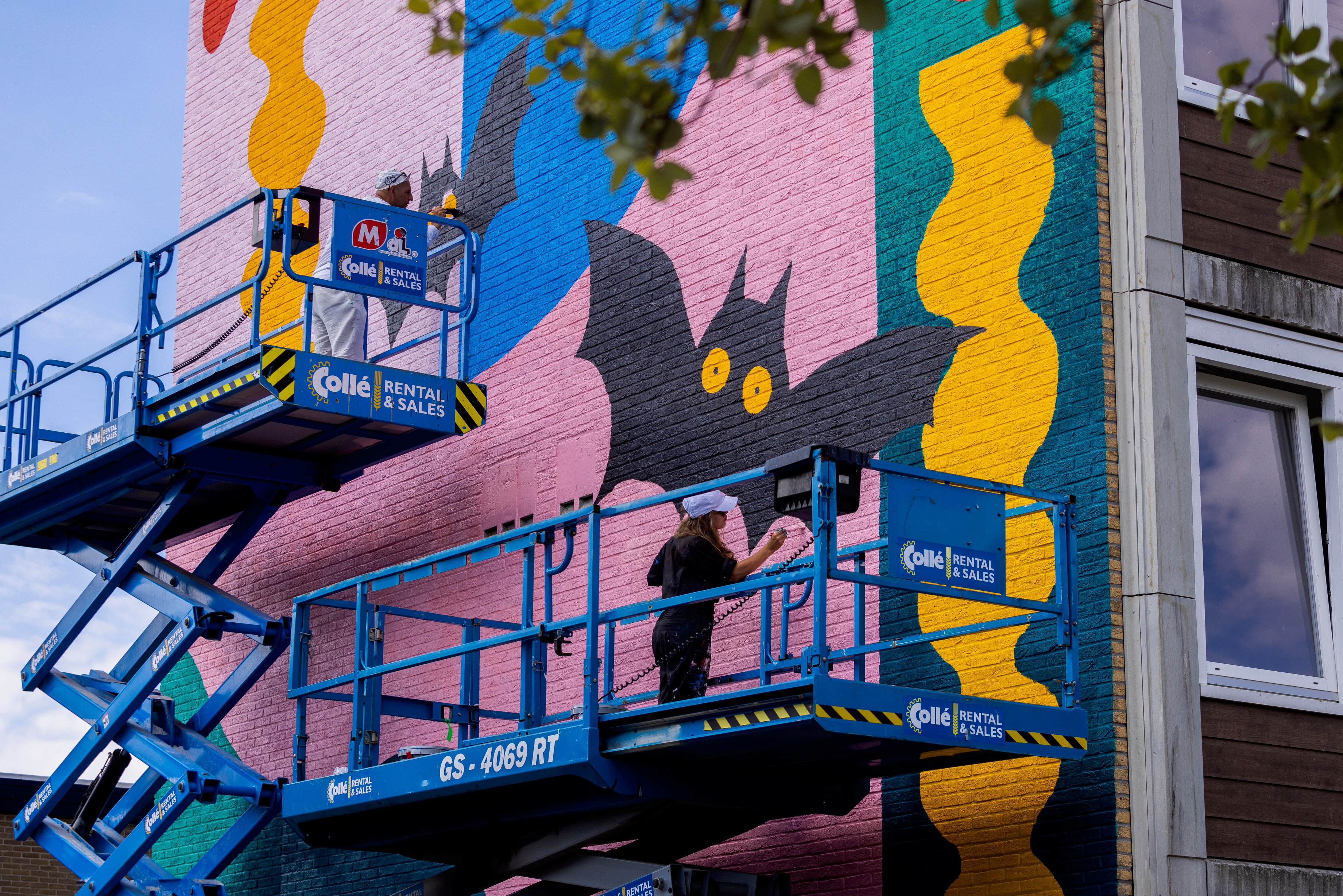 Twee hoogwerkers staan tegen de hoge muur van een flatgebouw af met daarop twee mensen (rechts vooraan de kunstenaar) die werken aan een grote muurschildering met kleurige vormen en een vleermuis 